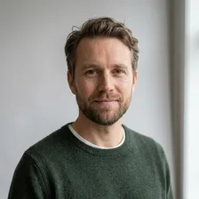 Eirik Sandvik smiler svakt mot kameraet, med kort bølgete hår, skjegg og grønn genser over hvit t-skjorte foran en lys vegg.